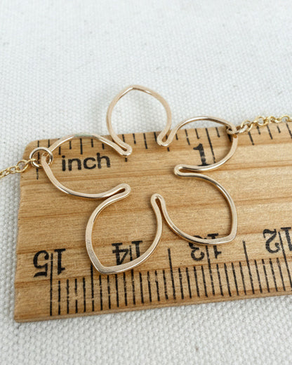 A close-up of a delicate gold necklace with a flower-shaped pendant, shown resting on a wooden ruler to indicate scale against a textured off-white fabric background.