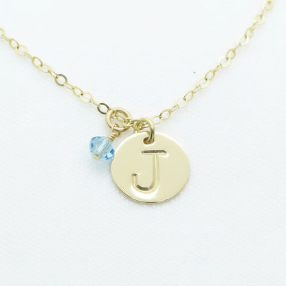 A gold necklace with a large initial letter 'J' accompanied by a small blue birthstone crystal, on a textured white background.
